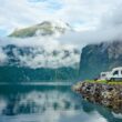 A row of motorhomes parked along an embankment of a turqoise lake and mountain range.