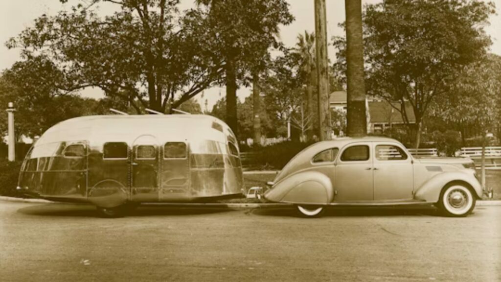 A photo of a small Airstream trailer from the 1930' hitched to a car and parked on the side of a road. 