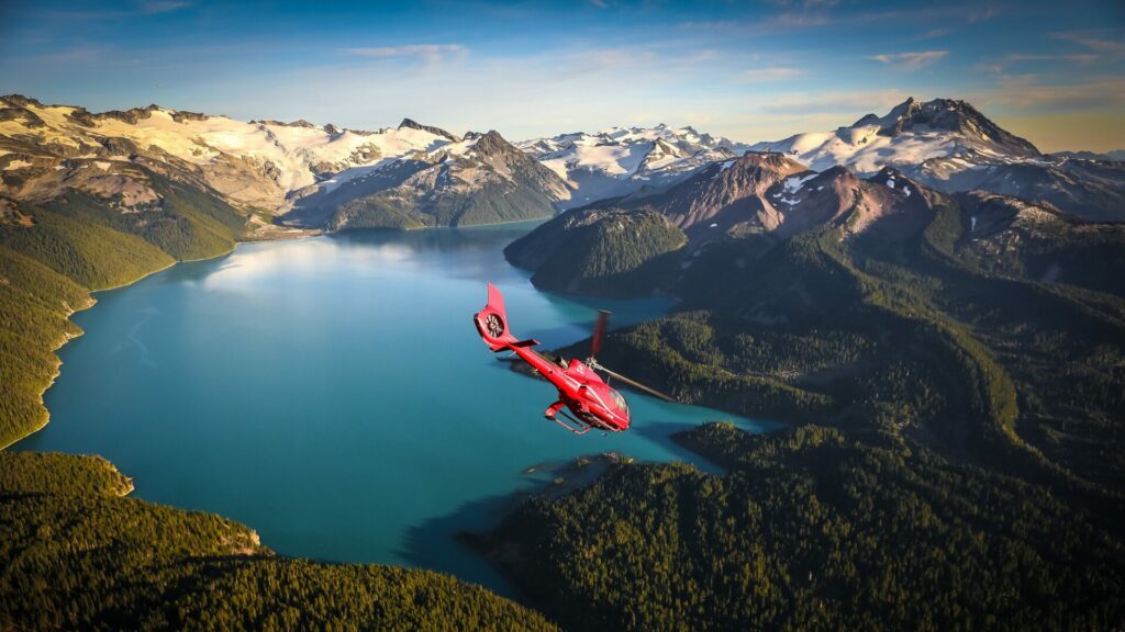 A photo of a red helicopter from BC Experience flying over turquoise waters surrounded by lush forests and snow-covered mountains. 