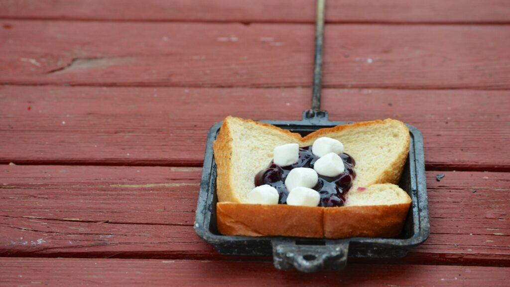 Half of a pie iron filled with a piece of white bread that has a pie filling with marshmallows atop it as it rests on a red picnic table. 