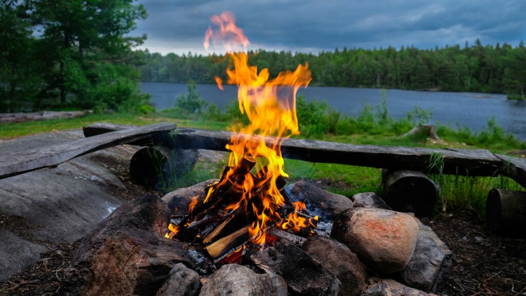 A campfire at dusk at a campground surrounded by logs with a body of water and lush dense green trees in the background.