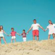 A photo of a family of five holding hands while standing on a sandy hilltop with a bright blue sky in the background.