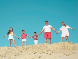 A photo of a family of five holding hands while standing on a sandy hilltop with a bright blue sky in the background.