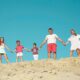 A photo of a family of five holding hands while standing on a sandy hilltop with a bright blue sky in the background.