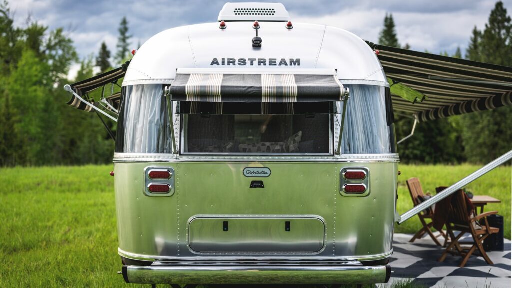 The Airstream Globtrotter trailer set up in a lush green site surrounded by dense forest. Beside it is a a little outdoor setup with two chairs, a table, and a rug. 