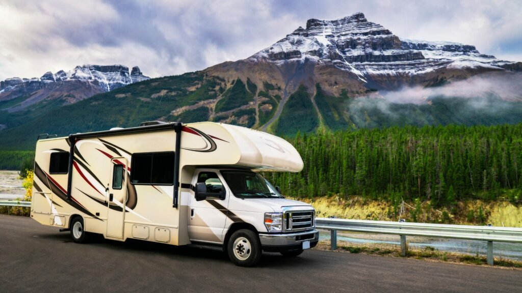 A motorhome parked in front of a guard rail on a freeway as a snow capped mountain stands tall behind it in the background.