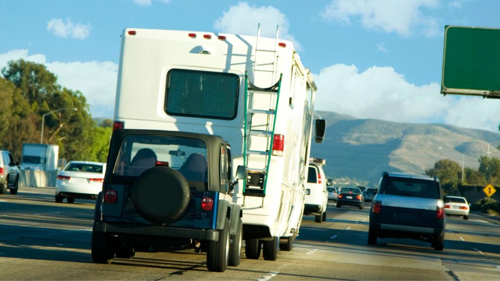 A blue jeep being flat towed behind a motorhome down a busy freeway.