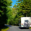 An RV driving down a heavily wooded road with bikes secured to the rear of it.