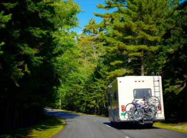 An RV driving down a heavily wooded road with bikes secured to the rear of it.