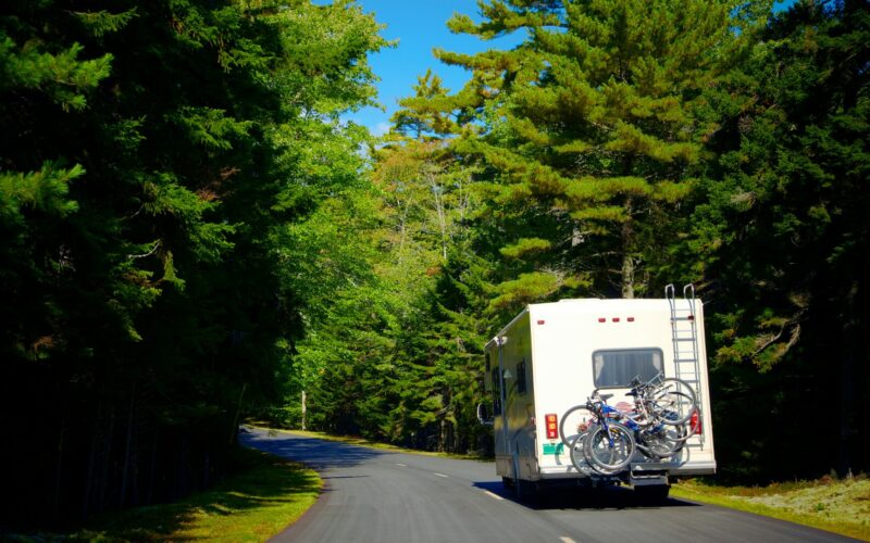 An RV driving down a heavily wooded road with bikes secured to the rear of it.