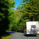 An RV driving down a heavily wooded road with bikes secured to the rear of it.