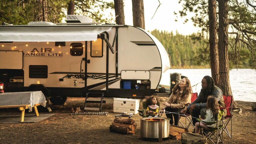 A small family of four sitting around a fire roasting marshmallows in front of a lake and their Thor RV trailer. 