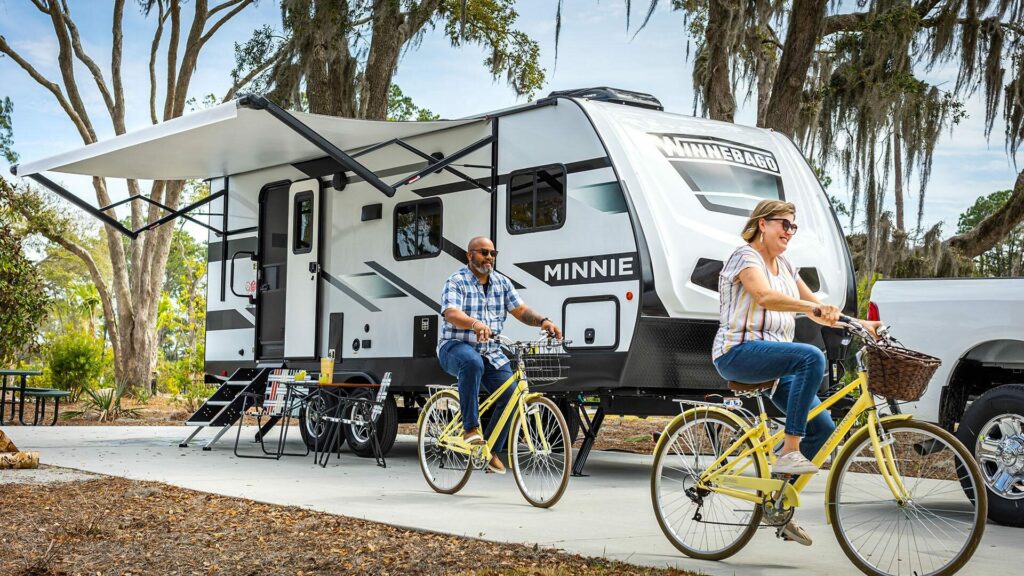 Two people riding yellow bikes beside their Winnebago travel trailer as it sits hitched on a white truck with the awning out. 