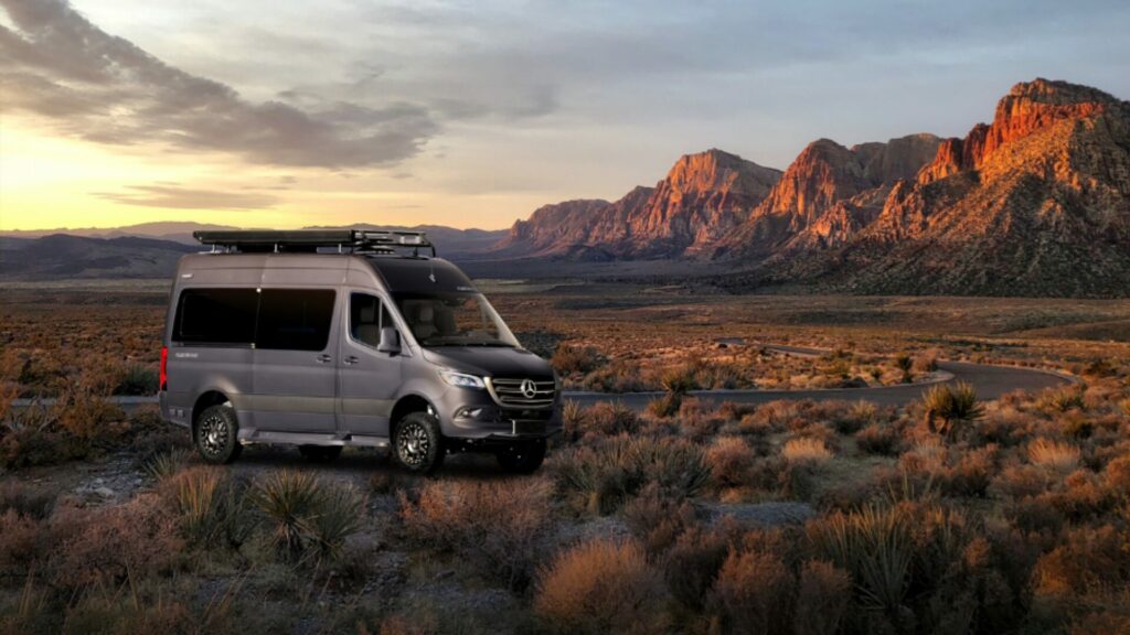 A grey Mercedes Travel Van from one of the largest RV Manufacturers, the REV Group. It's sitting in front of a red rocky cliffs beside a winding road. 