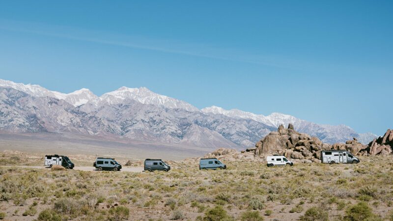 A lineup of six Winnebago travel vans in front of a mountain range.