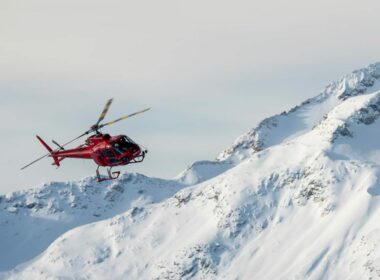 An aerial photo of a red Whistler Helicopter Tour Helicopter flying over snowy mountains.