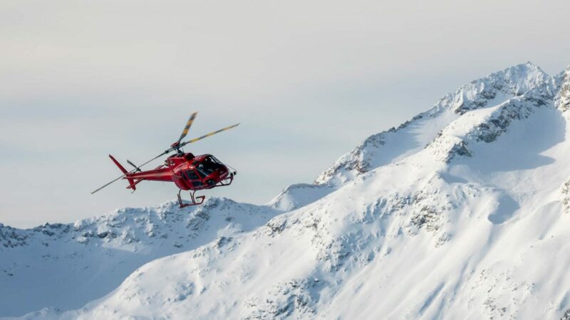 An aerial photo of a red Whistler Helicopter Tour Helicopter flying over snowy mountains.