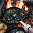 Person holding a cast iron pan over open flames as two other pans sit resting atop the campfire.