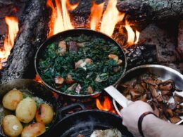 Person holding a cast iron pan over open flames as two other pans sit resting atop the campfire.