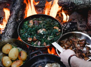 Person holding a cast iron pan over open flames as two other pans sit resting atop the campfire.