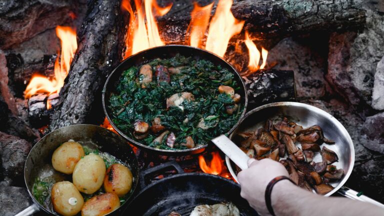 Person holding a cast iron pan over open flames as two other pans sit resting atop the campfire.