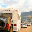 A red Jeep being towed behind a motorhome on a freeway with blue cloudy skies and rolling hills in the background.