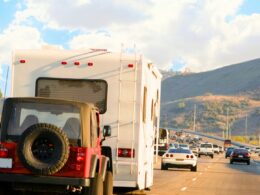 A red Jeep being towed behind a motorhome on a freeway with blue cloudy skies and rolling hills in the background.