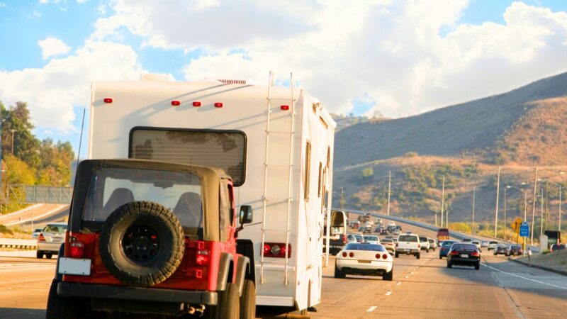 A red Jeep being towed behind a motorhome on a freeway with blue cloudy skies and rolling hills in the background.