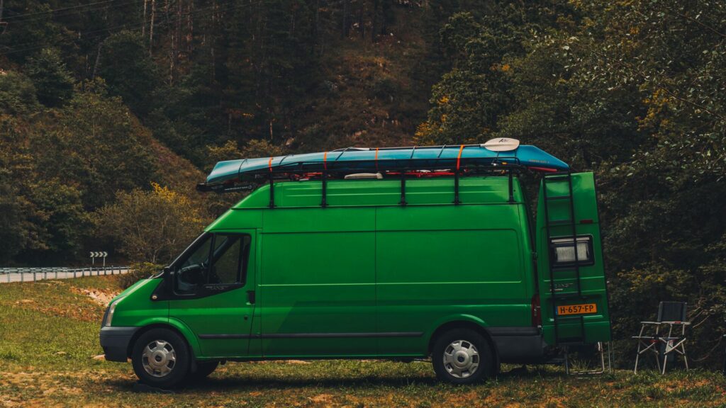 A converted camper van in a vibrant kelly green parked with lawn chairs behind it and water sports vessels attached to the top of it. 