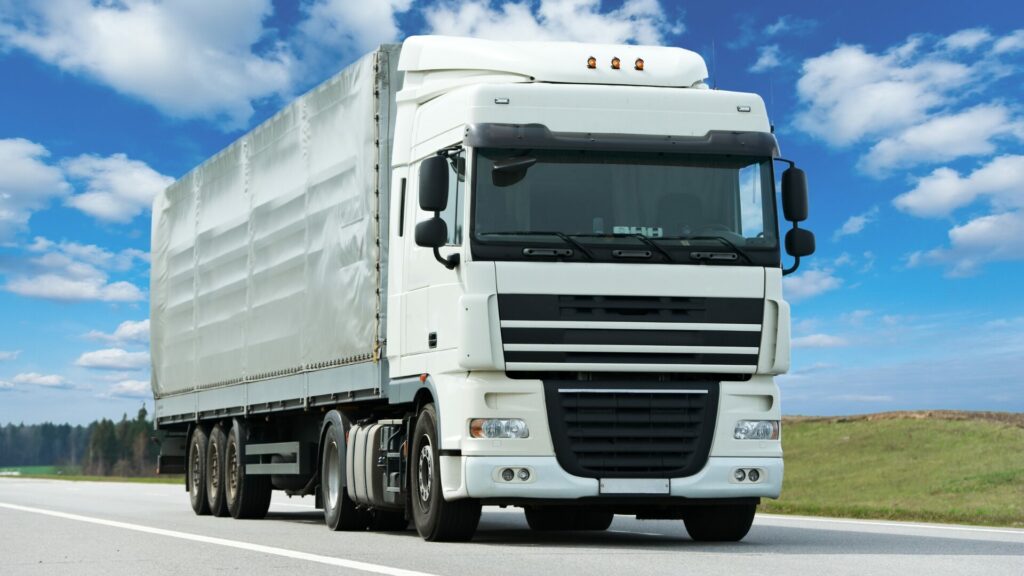 A articulated lorry driving on a highway with a bright blue sky and green plains behind it. 