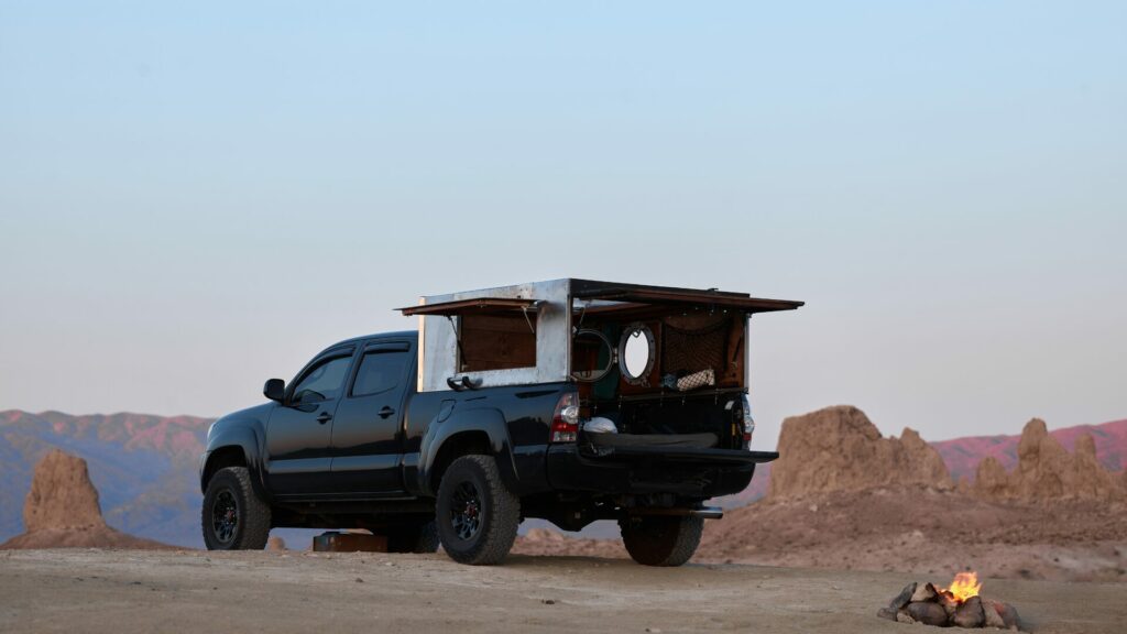 A truck with a converted bed for camping in. A small firepit is built and lit beside it in a desert rocky surrounding. 