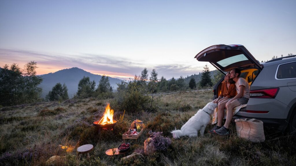 A couple and their large white dog sitting in the back of their SUV in front of a picnic set up beside a firept while the sun sets behind a large forested peak. 