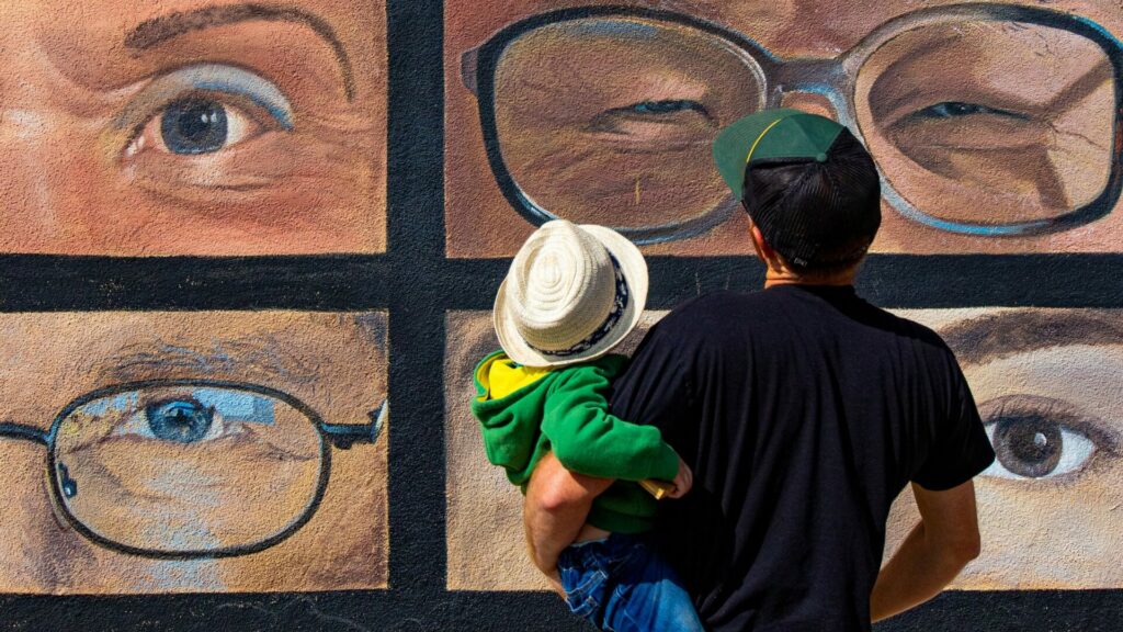 A person holding a child while looking at a mural of many different eyes. 