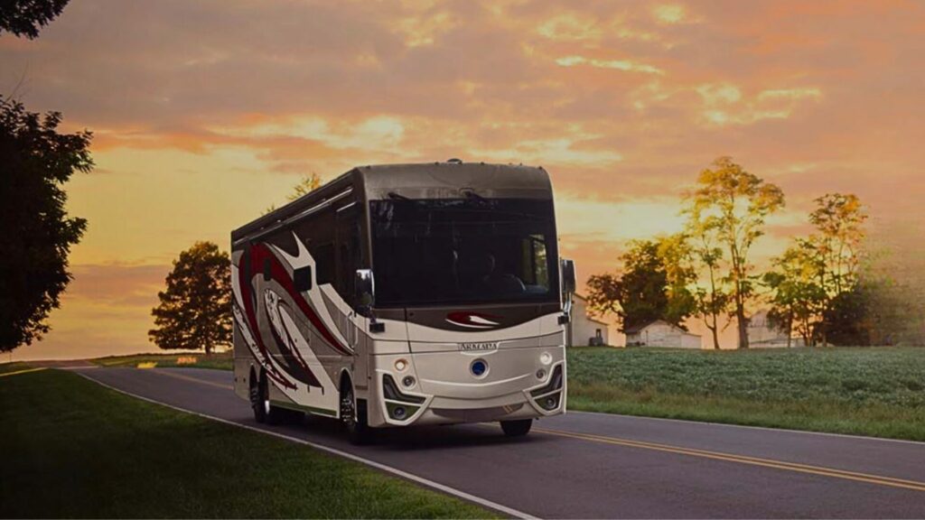 A motorhome from Holiday Rambler driving down a road with a setting sun behind it as it paints the sky orange. 