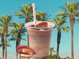 A shake with dates upon and beside it as it sits in front of a photo of date trees side by side with a bright blue background.